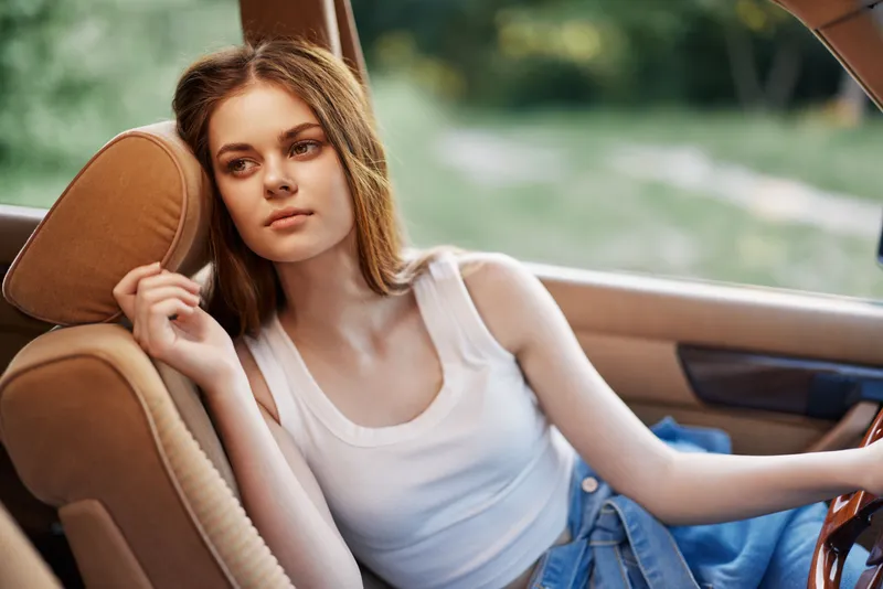 Young woman with long brown hair in summer outfit, relaxing in car, expressing calm emotions in natural outdoor setting, showcasing vibrant colors and seasonal vibes.