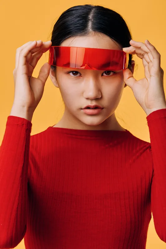 Young woman with serious expression wearing red shirt adjusting futuristic red visor on orange background. Studio portrait with modern fashion style and clean composition in closeup.
