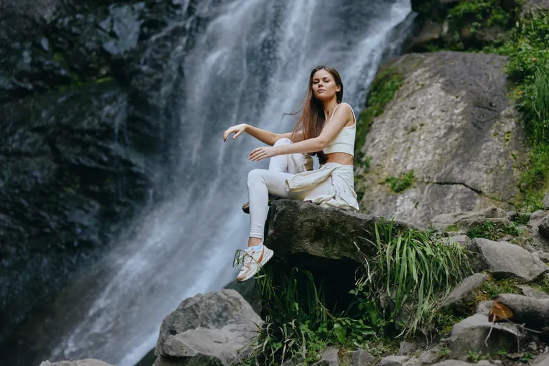 woman sitting by waterfall wearing white clothes poses on rocks surrounded by lush greenery and natural outdoor scenery in daylight