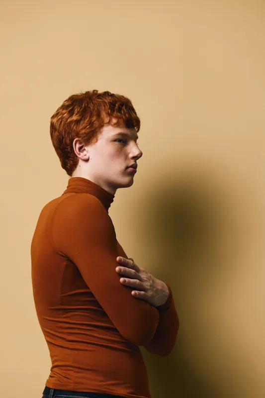 Young man with red hair in brown turtleneck sweater standing in profile with crossed arms on beige background. Casual style, confident pose, isolated portrait.