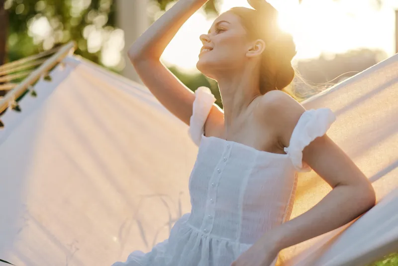 Young woman relaxing in a hammock, wearing a light summer dress, enjoying the sunset with a serene expression, surrounded by greenery and warm sunlight