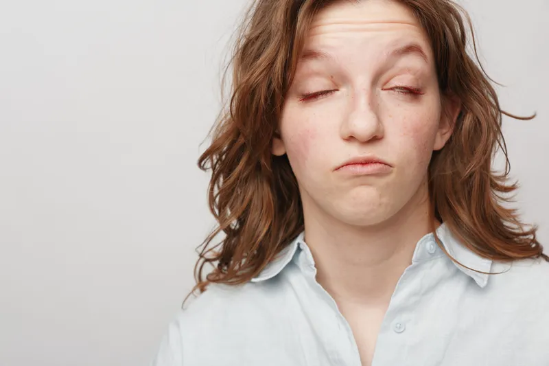 Young woman with messy hair and closed eyes makes a funny unimpressed face wearing a light shirt on plain background. Concept of boredom, frustration, and casual lifestyle.