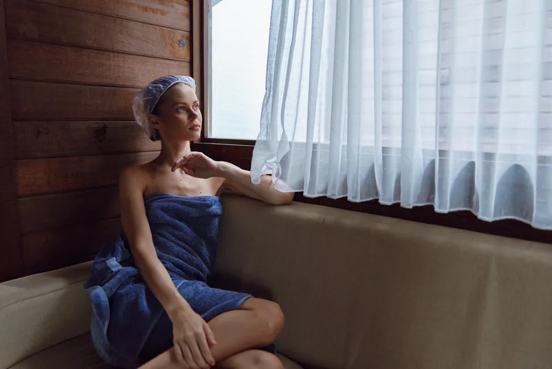 Relaxed woman wrapped in a towel sits by the window in a wooden sauna room, wearing a hair cap and enjoying soft natural light and peaceful ambiance indoors.