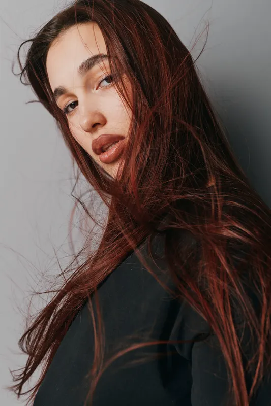 Woman with long red hair and natural makeup posing against a gray background. She wears a black top with hair flowing and looks confidently at the camera in studio lighting.