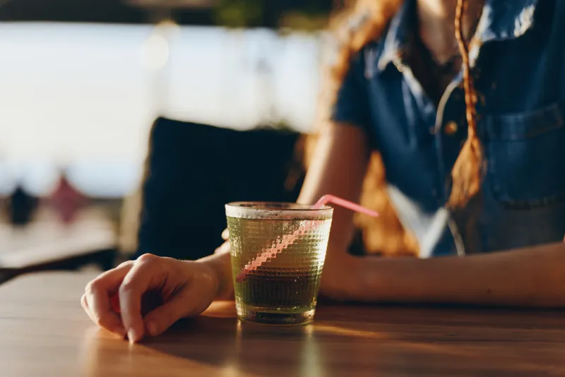lifestyle woman in modern boho-western denim street style holding a glass with a straw, warm natural light and film color tones creating relaxed atmosphere
