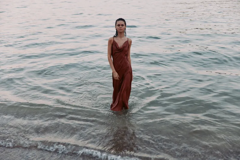 Solitary woman stands waist-deep in a calm sea, wearing a flowing red dress as gentle waves lap the shore, creating a serene, cinematic moment of solitude.