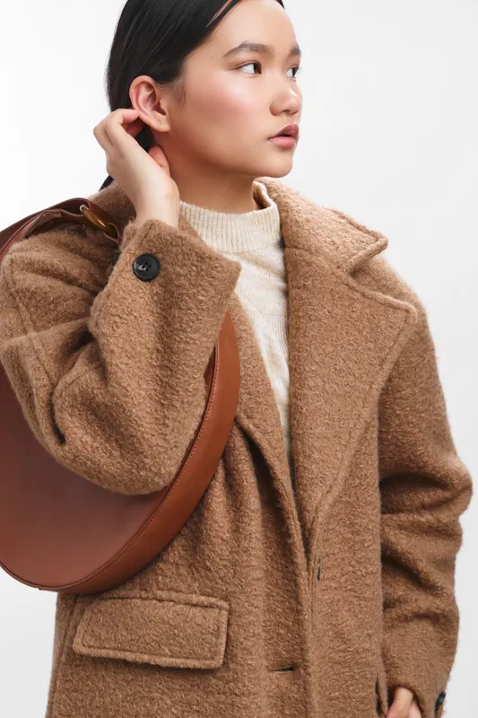Asian woman wearing a warm brown coat stands in a bright studio, adjusting her hair and looking to the side, conveying confidence, fashion, and poised urban style. Her expression is calm and