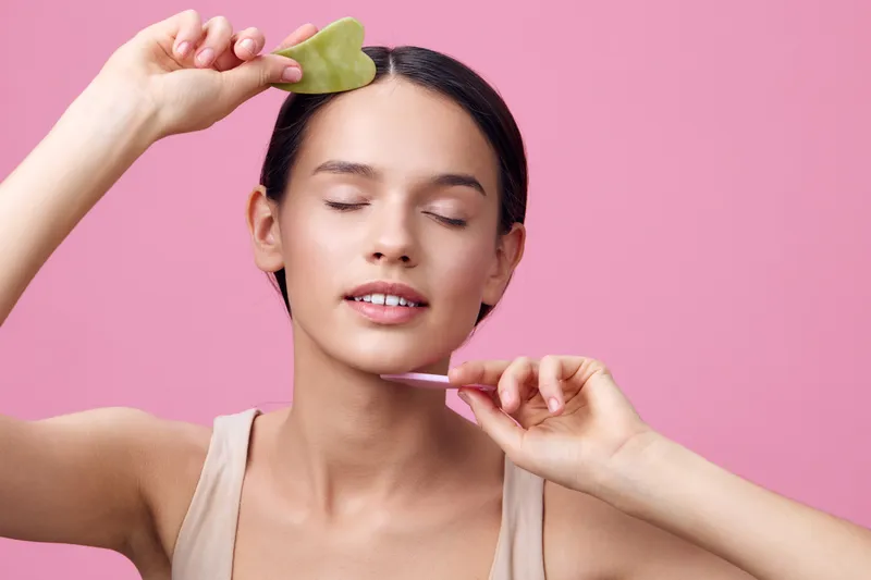 Beautiful young model using a green jade gua sha for facial massage, promoting skincare and wellness, with a soft pink background