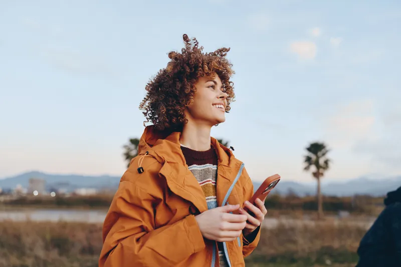 Woman lifestyle portrait outdoors, smiling with curly hair in an orange jacket, holding a smartphone and looking at the screen, warm sunset light across fields, travel mood, nature scene.