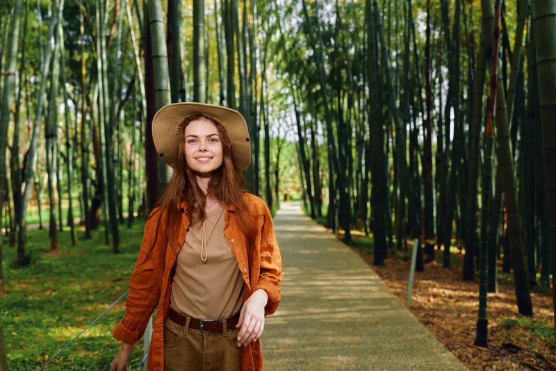 Woman walking in bamboo grove on a paved path, smiling portrait with straw hat and autumn coat. Nature trail scene, casual fashion, leisure outdoors and peaceful green surroundings.