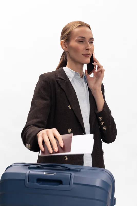 Businesswoman in dark blazer holds passport and boarding pass while speaking on smartphone. Blue suitcase in foreground. Concept of travel, business trip, and modern professional lifestyle.