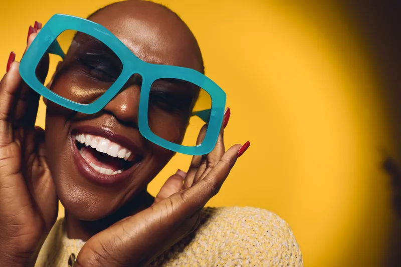 Happy smiling black woman with dark skin and bald head wears oversized blue glasses on yellow background. Concept of joy, fashion, beauty, and confident African ethnicity female model.