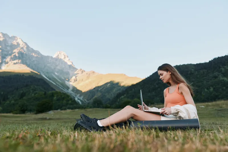 A young woman sits on a grassy field with a laptop, enjoying a sunny day in a mountainous landscape, creating a calm and productive outdoor workspace.