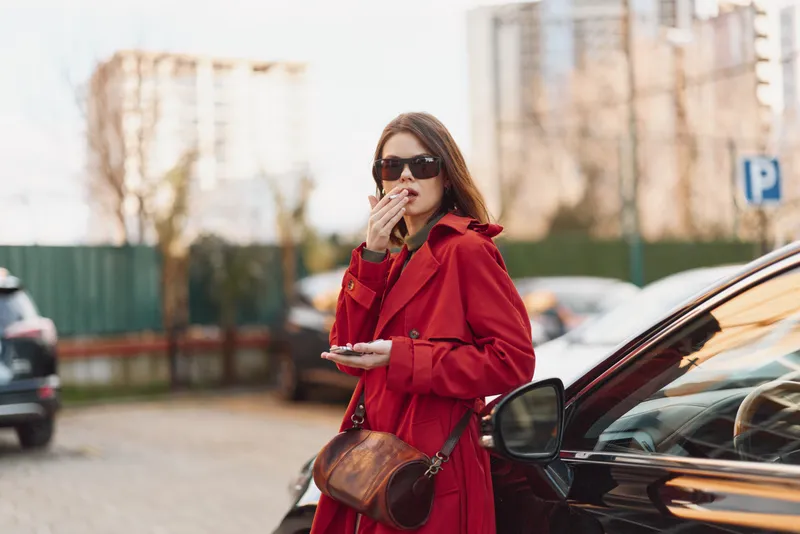 Stylish woman in a red coat and sunglasses stands beside a car in an urban parking lot, smoking calmly, conveying bold street fashion and confident attitude