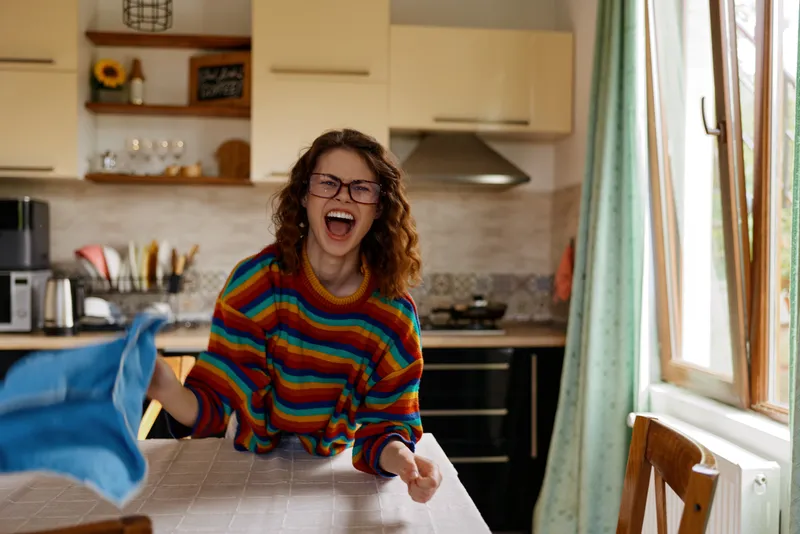 Cheerful woman with curly hair and glasses laughs in a bright home kitchen, wearing a colorful striped sweater while wiping a table with a blue cloth during a casual cleanup at home