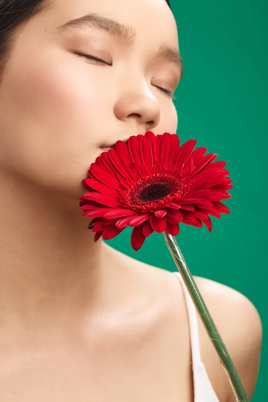 asian woman with a red flower held near her face, a soft serene portrait under bright studio light, emphasizing natural beauty and calm mood.