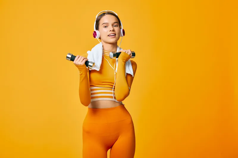 Athletic woman fitness portrait in matching orange activewear lifting small dumbbells with a towel around her neck, wearing headphones and a confident smile on a vivid orange background.