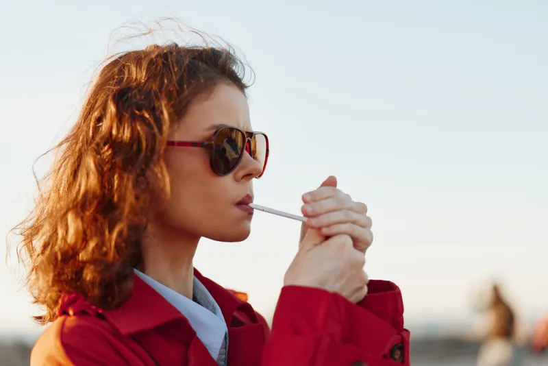 Young woman with curly red hair wearing a red jacket and sunglasses lights a cigarette outdoors in warm soft light, creating a calm confident mood as the sun sets.