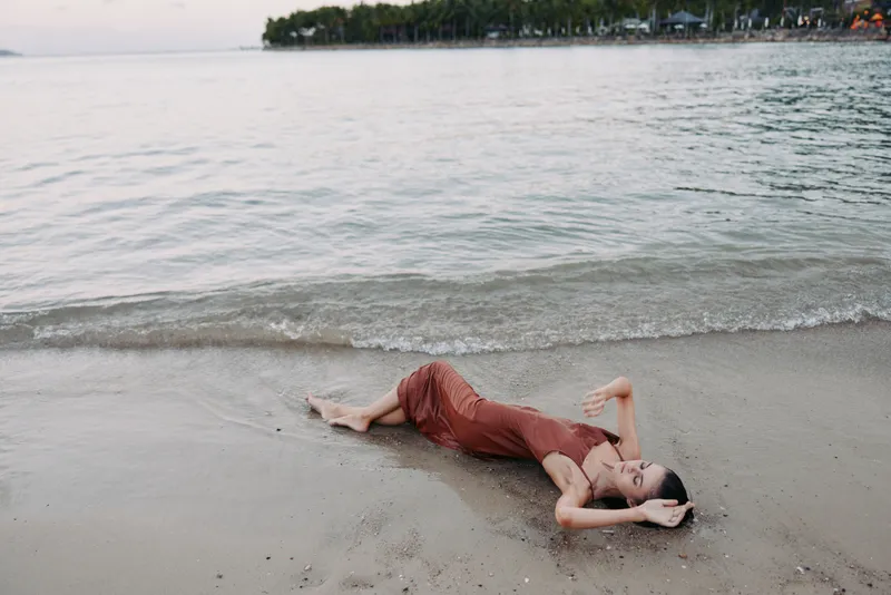 A solo figure rests on wet sand near calm shoreline, fabric dress melting into the tide as soft light paints a tranquil seaside moment with gentle waves and distant land and silence.