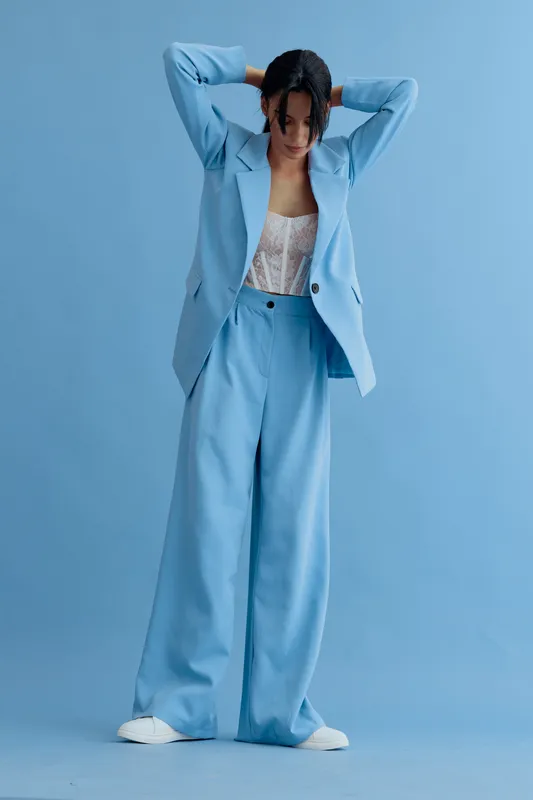 Woman in blue suit posing against a matching blue background, wearing white sneakers and a lace top, showcasing modern fashion in minimalist style.