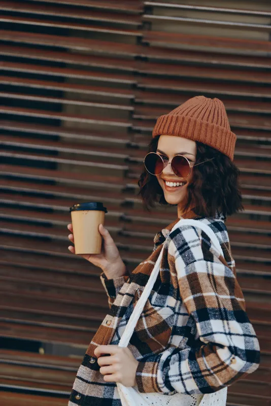 Woman holding coffee, smile and sunglasses in urban street, beanie and plaid jacket exuding authenticity and candid lifestyle with golden hour glow, mindful living and emotional storytelling.