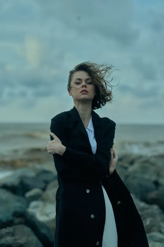Woman in a long dark coat stands on a rocky shore, windblown hair, stormy sky, fashion at the sea with a moody coastal atmosphere.