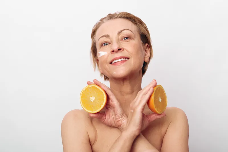 Smiling middle aged woman with natural skin holds two orange halves near face with cream on cheek. Concept of skincare, healthy lifestyle, natural beauty, wellness, and citrus freshness.