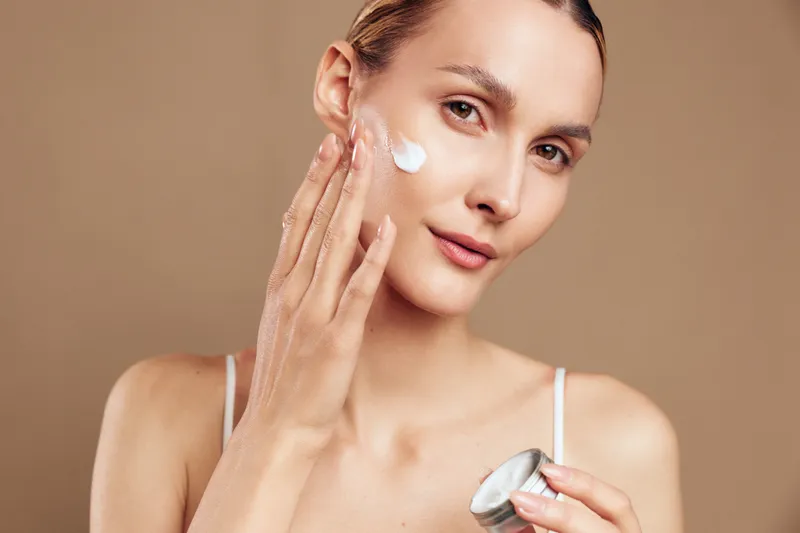 A young woman applies moisturizer to her face in a softly lit, neutral-toned setting, conveying a calm skincare routine and a moment of personal self care.