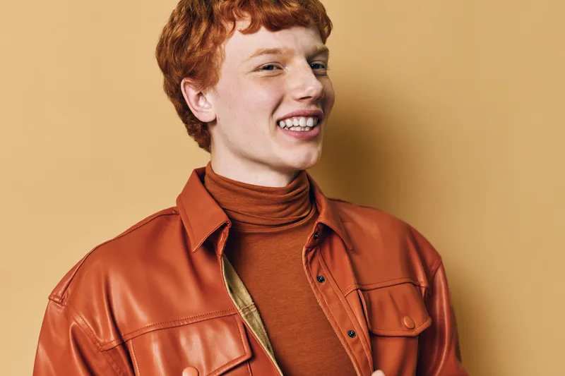 Young man with red hair smiles wearing a brown turtleneck and orange leather jacket against beige background. Casual style with trendy autumn outfit and confident expression.