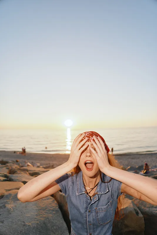 surprised woman covering eyes with hands at the beach during sunset with calm sea and clear sky in the background