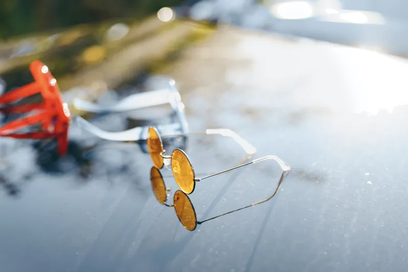 An outdoor scene features a pair of sunglasses resting on a glossy surface under bright daylight, creating a casual mood with vivid reflections and soft background details.