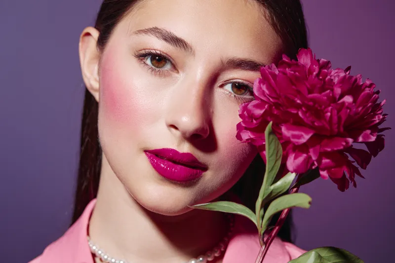 Close up portrait of young woman with creative vibrant pink makeup holding bright pink flower. Studio shot with purple background showcasing beauty fashion concept.