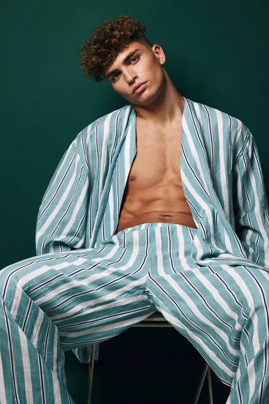 Young man with curly hair wearing striped loose pants and open shirt sitting on chair against dark background with serious expression and relaxed posture.