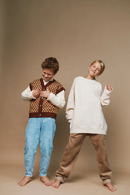 Two joyful children pose playfully against a neutral backdrop, showcasing cozy casual outfits and carefree movement, capturing a warm moment of friendship and creative style.