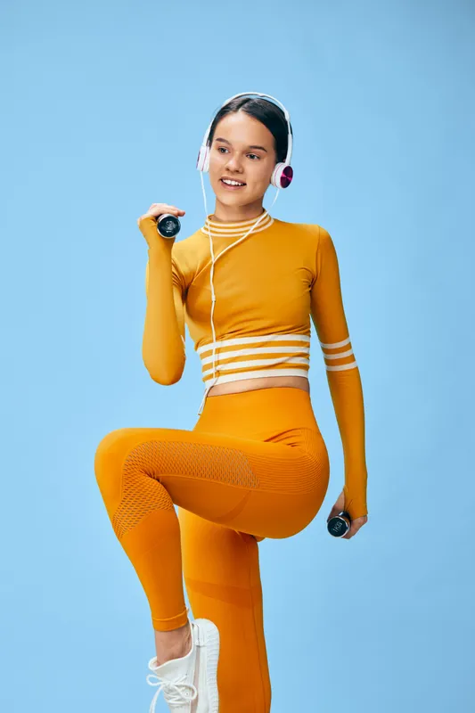 Woman fitness athlete in an orange athletic outfit lifts dumbbells, wearing headphones, mid-step against a blue backdrop, showcasing energy, movement, and focus during an intense workout session.