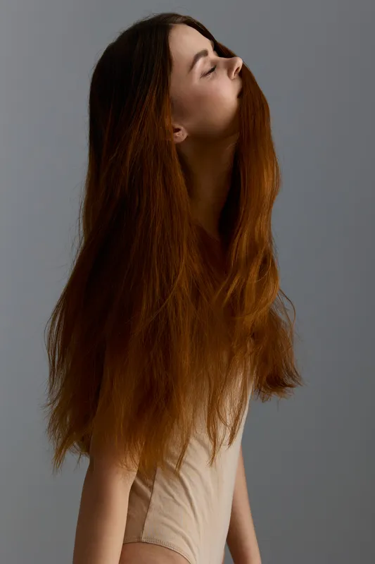 fashion woman with long red hair posing with closed eyes in beige bodysuit against gray background, showcasing natural beauty and elegance in studio lighting environment
