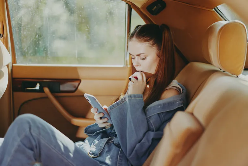 Young woman with long hair in denim outfit sitting in a car, focused on her smartphone, showcasing a relaxed mood and warm colors.