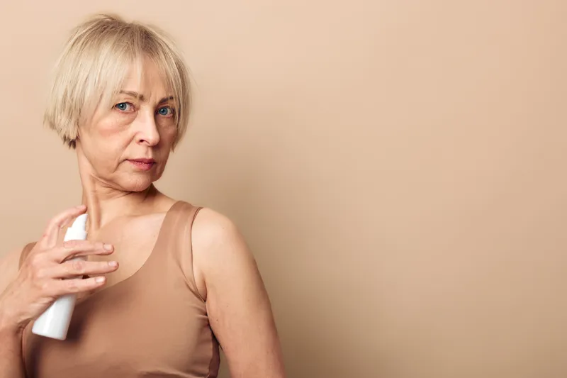Mature woman with short blonde hair applies deodorant to her underarm wearing beige sleeveless top. Neutral background highlights personal hygiene and skincare routine concept.