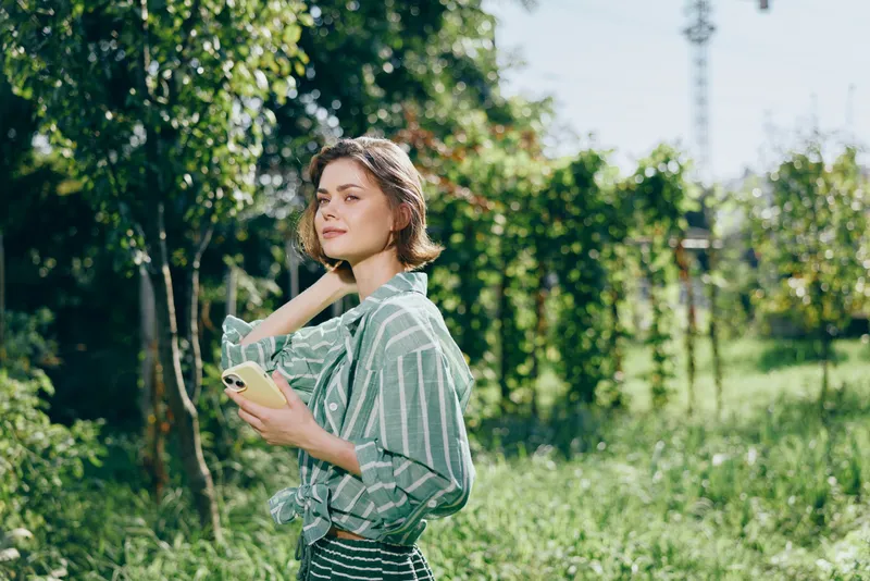 A portrait of a young woman standing in a sunlit garden, looking off to the side while holding a phone.