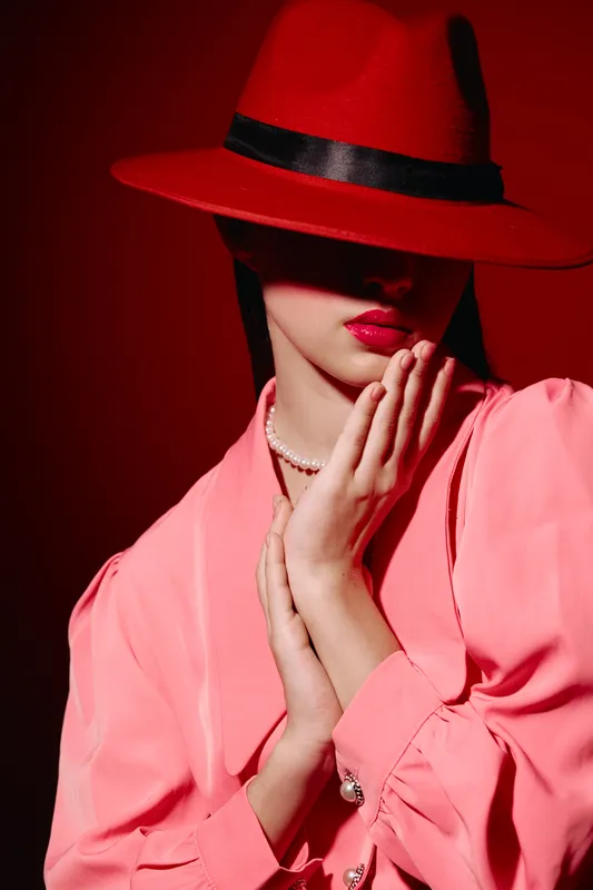 Fashion model in pink blouse and red hat poses against matching red background, lips painted bold red, wearing pearl necklace creating glamor and elegance beauty concept.