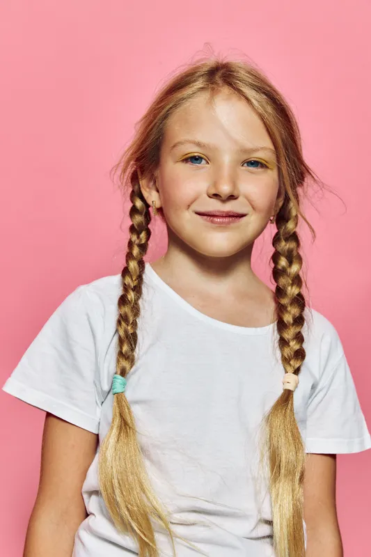 Smiling young girl with long blonde hair in two braids wearing a white t-shirt standing against a pink background.