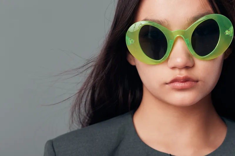 Young woman with long dark hair wearing large green sunglasses and dark outfit posing against plain gray background. Closeup portrait with stylish accessory and serious expression.
