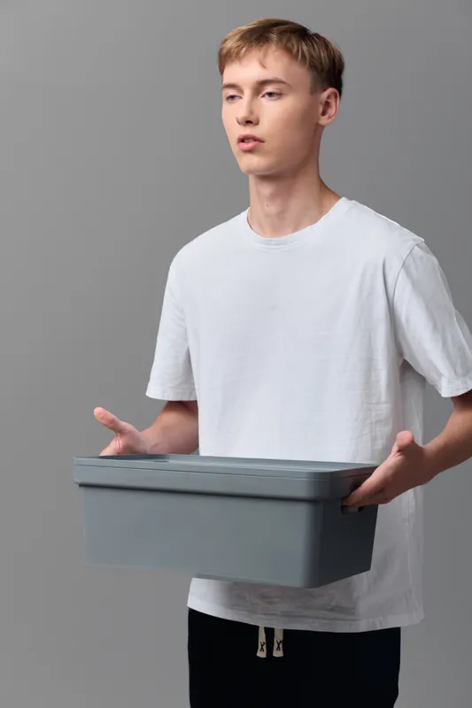 Young man in a white t shirt holds a gray storage box, illustrating packaging and logistics in a clean studio setting, with emphasis on shipment and simple order fulfillment.