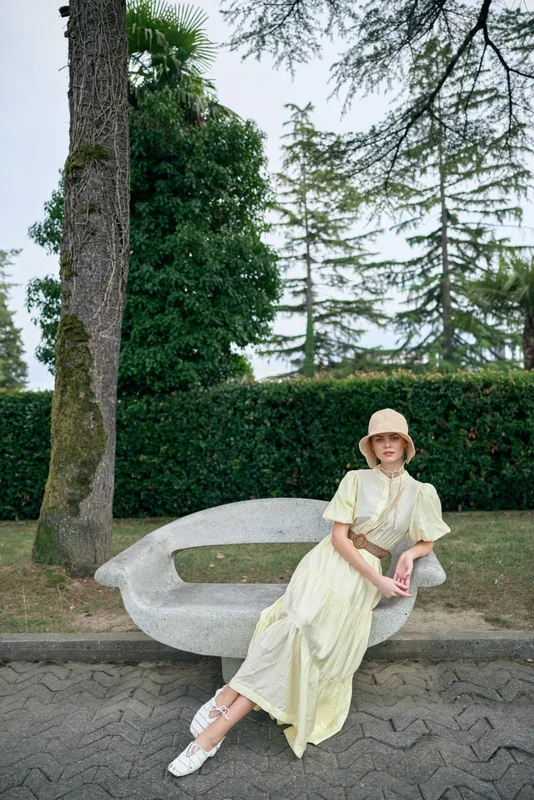 Woman sitting on sculptural bench in park wearing flowing dress and hat portrait conveying authenticity and inclusivity relaxed pose among trees and curated landscape