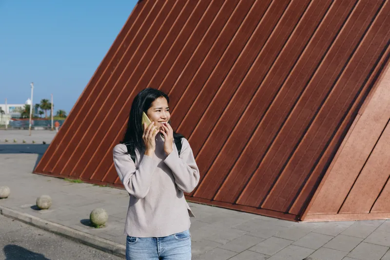 A woman talks on her phone outdoors beside a bold red architectural wall, wearing casual clothes under bright sunlight, capturing an urban street scene with distant palm trees