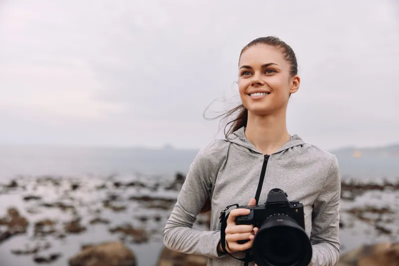 A smiling young woman with long hair in a gray sports hoodie holds a camera, enjoying her time by the rocky shoreline at dusk, embodying passion for photography