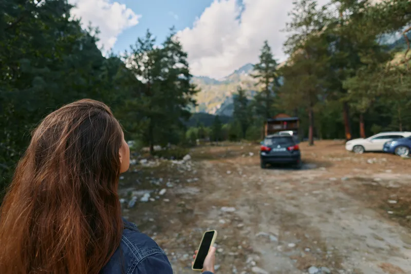 A person with long hair stands in a rustic parking area beside a forest, looking at a smartphone with a calm, focused expression, surrounded by trees and distant peaks.