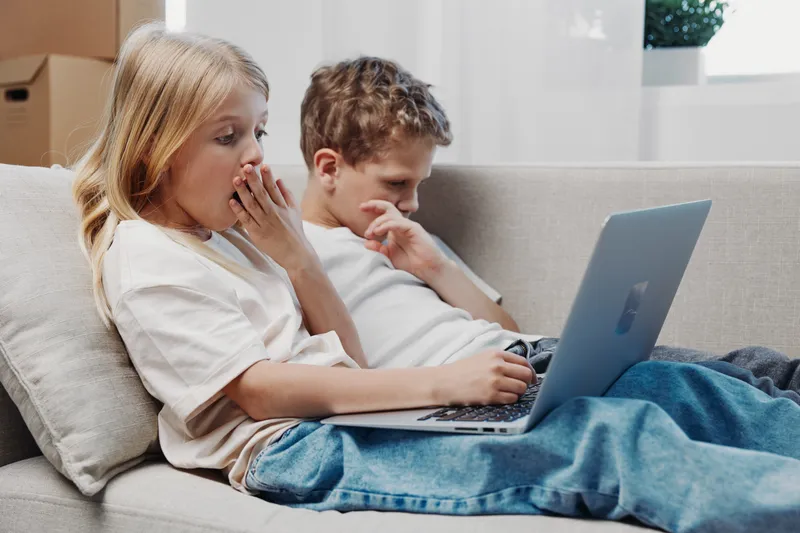 Two children sitting on sofa using laptop, one yawning and the other focused. Casual home environment, concept of childhood boredom and digital interaction.