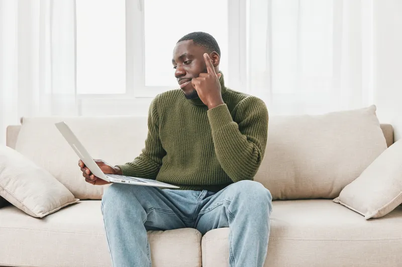Thoughtful young man in a cozy sweater using a laptop at home, reflecting on ideas and enjoying a relaxed atmosphere with natural light