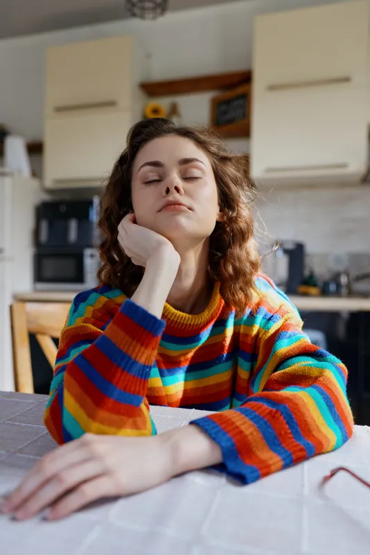 A young woman with a rainbow striped sweater rests her chin on her hand at a bright kitchen table, eyes closed, conveying a calm, daydreaming moment in a cozy home setting.